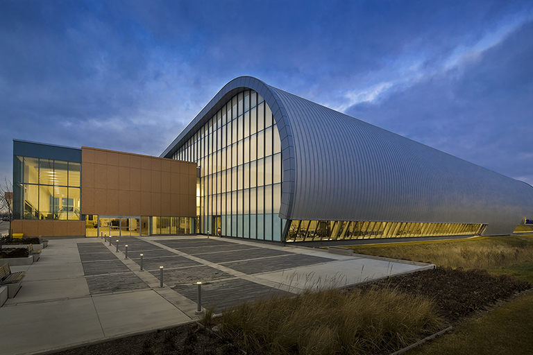 Large, curved, metal-clad structure with curtain wall windows adjoining a rectangular metal and glass building.